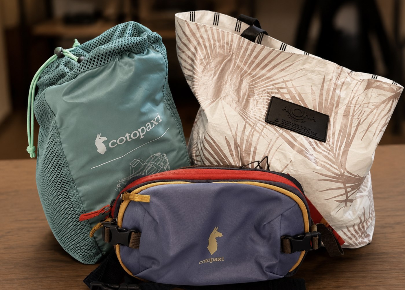 A Cotopaxi stuff sack, hip pack and a palm-print Aloha tote on a table