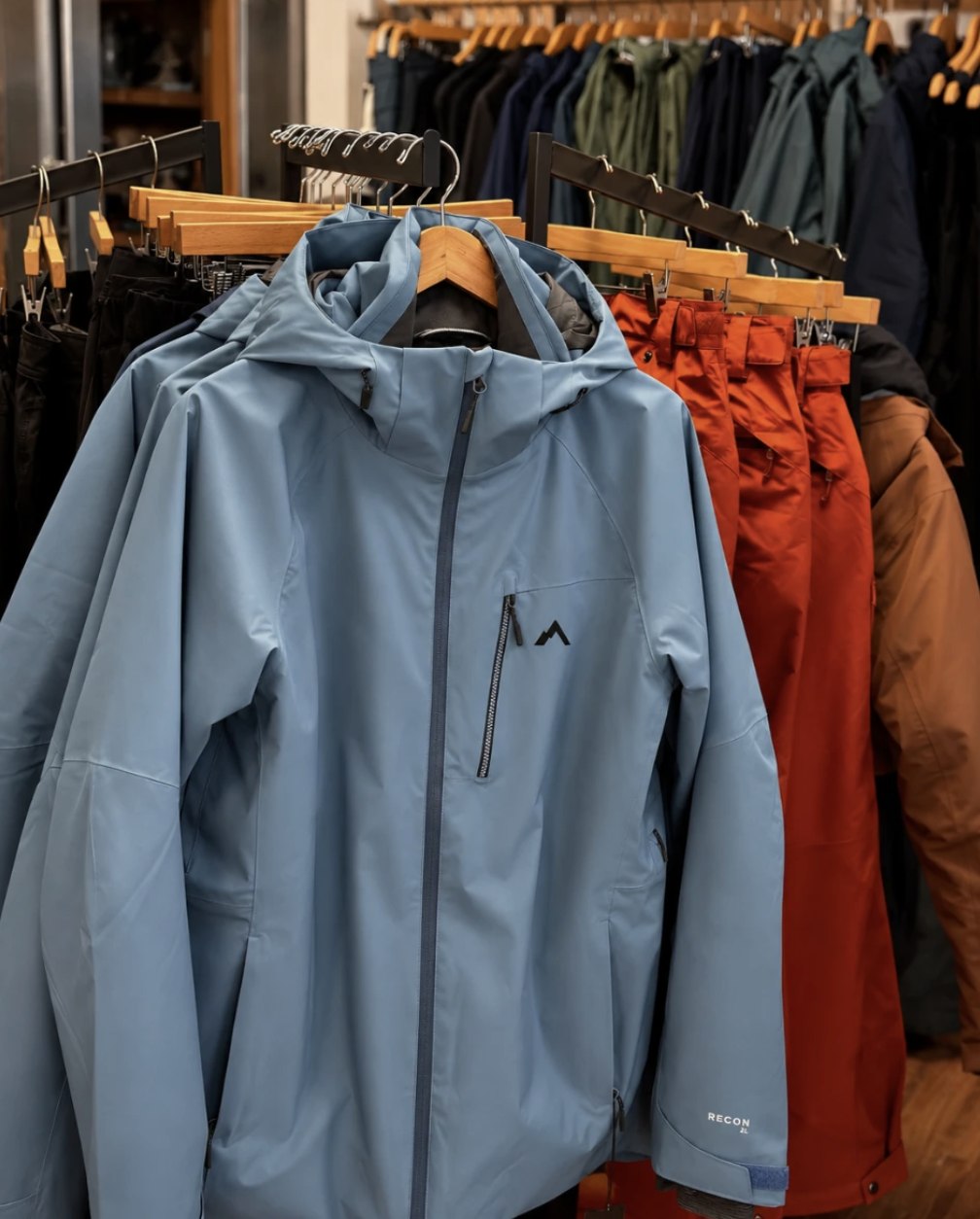 A blue Strafe RECON insulated shell jacket on a rack beside orange snow pants